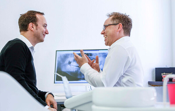 Zahnarzt berät Patienten freundlich in moderner Praxis mit Röntgenbild im Hintergrund.