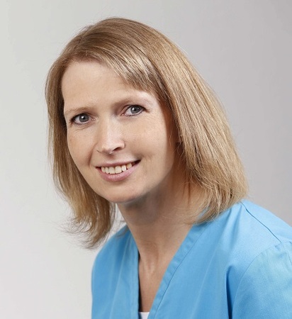 Porträt einer lächelnden Frau in medizinischer Berufskleidung vor neutralem Hintergrund.