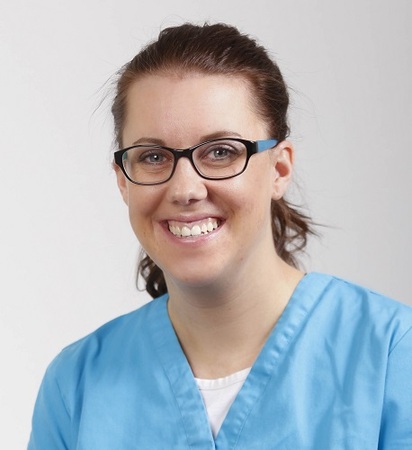 Portrait einer jungen Pflegekraft mit Brille in blauer Arbeitskleidung, freundlich lächelnd.